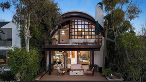 Una casa de dos pisos con balcón, un patio con mobiliario exterior y un amplio ventanal con vistas al jardín.