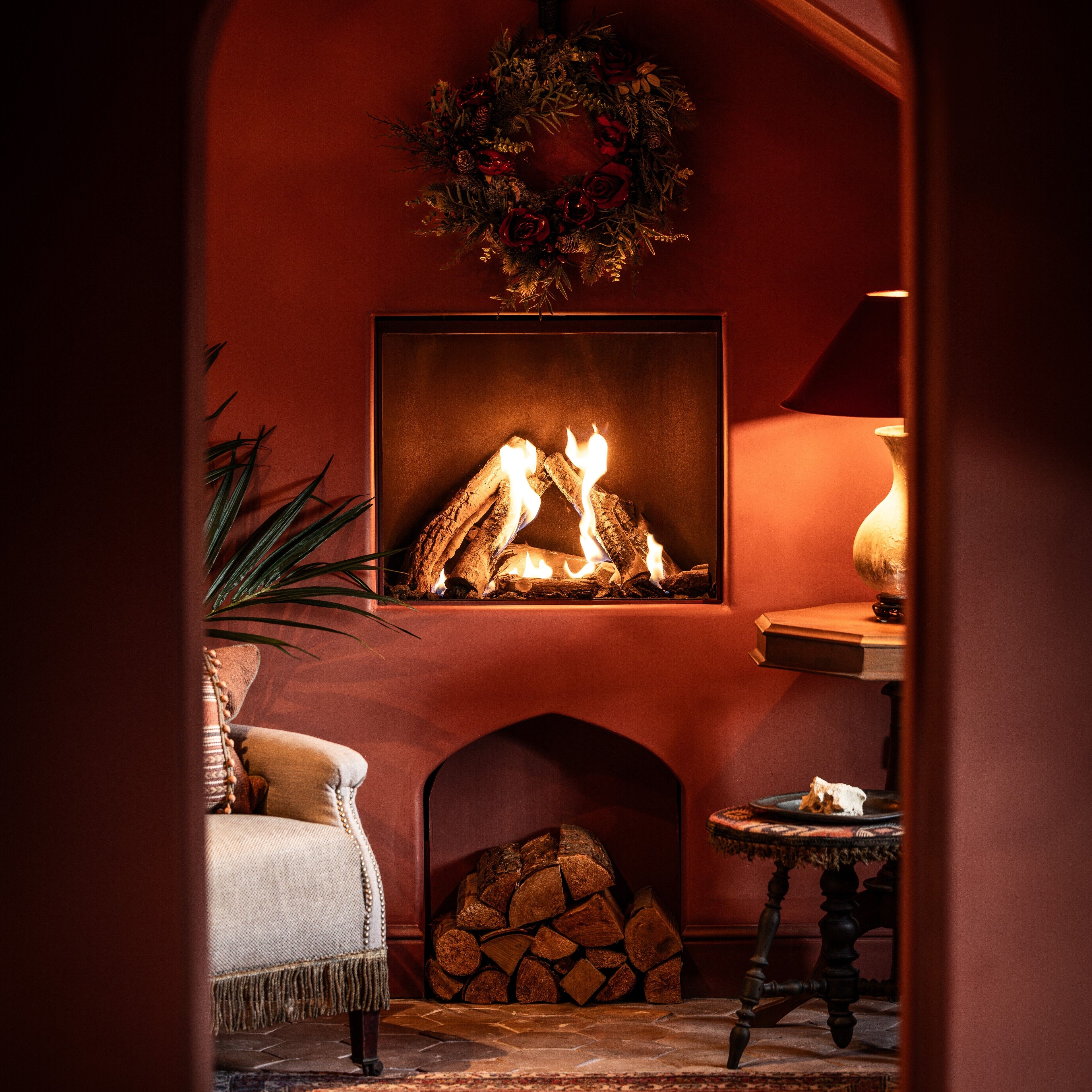 A cozy fireplace with a log fire, a mantel with a wreath, and a comfortable seating area with a chair and a small table.