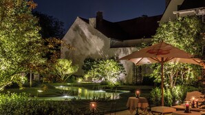 An outdoor patio area with a pool, tables, and chairs, surrounded by lush greenery and a stone wall. - The Notary (Bruges)