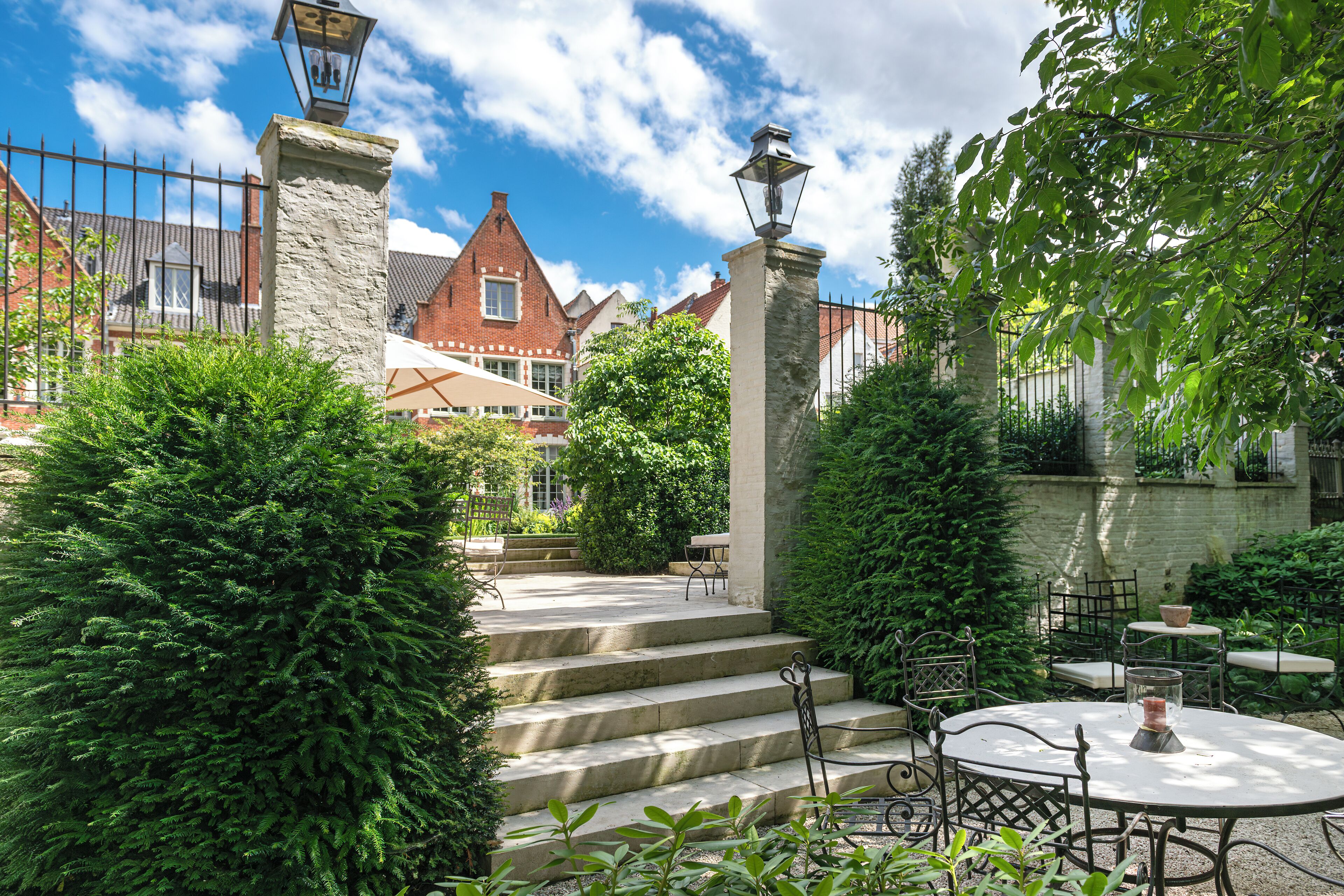 A courtyard with stone steps leading to a table and chairs, surrounded by greenery and a brick building in the background.