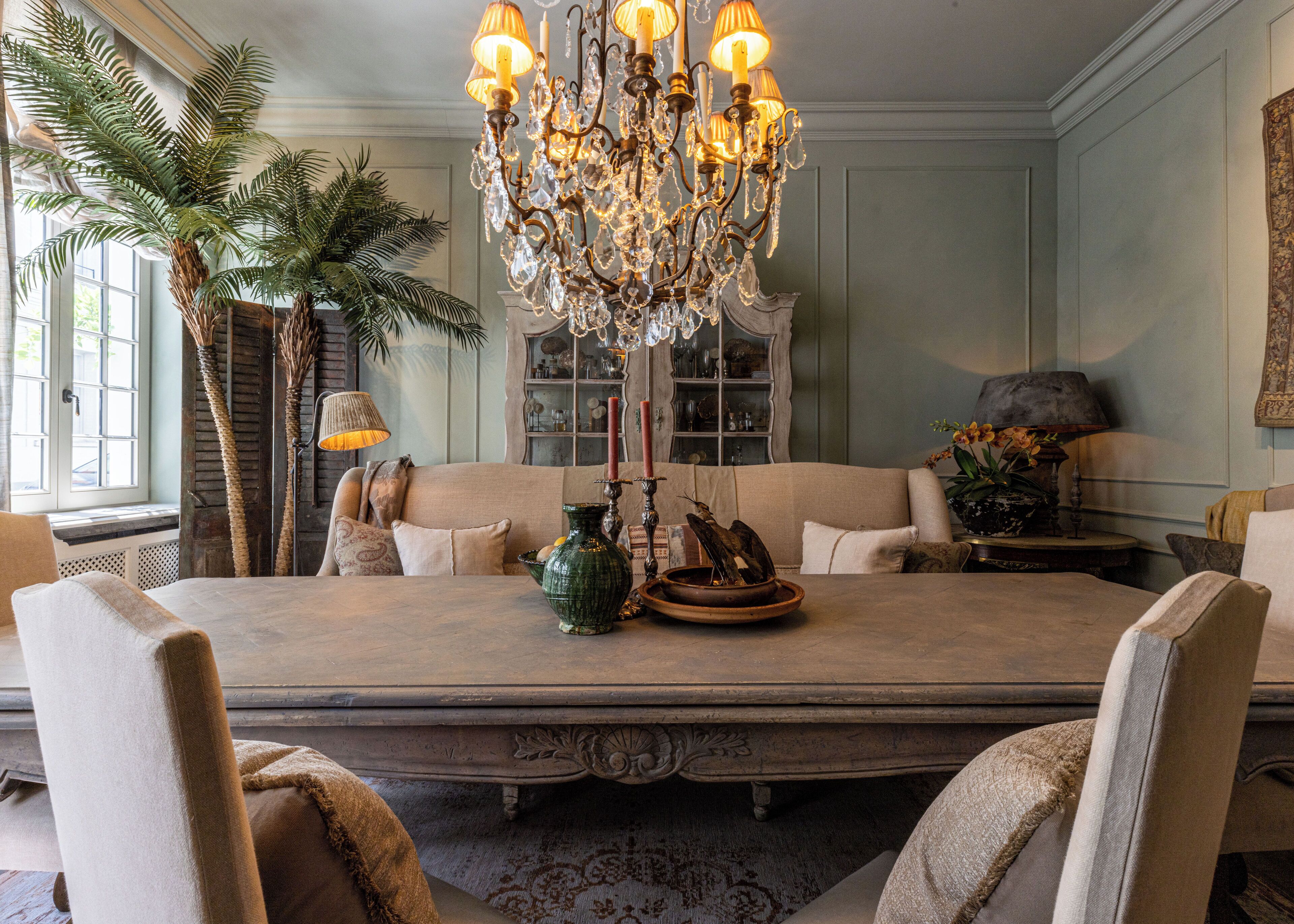 A dining room with a large table, chairs, a chandelier, a potted plant, and a bookshelf.