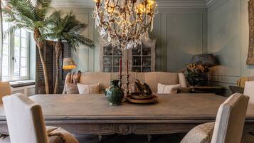 A dining room with a large table, chairs, a chandelier, a potted plant, and a bookshelf.