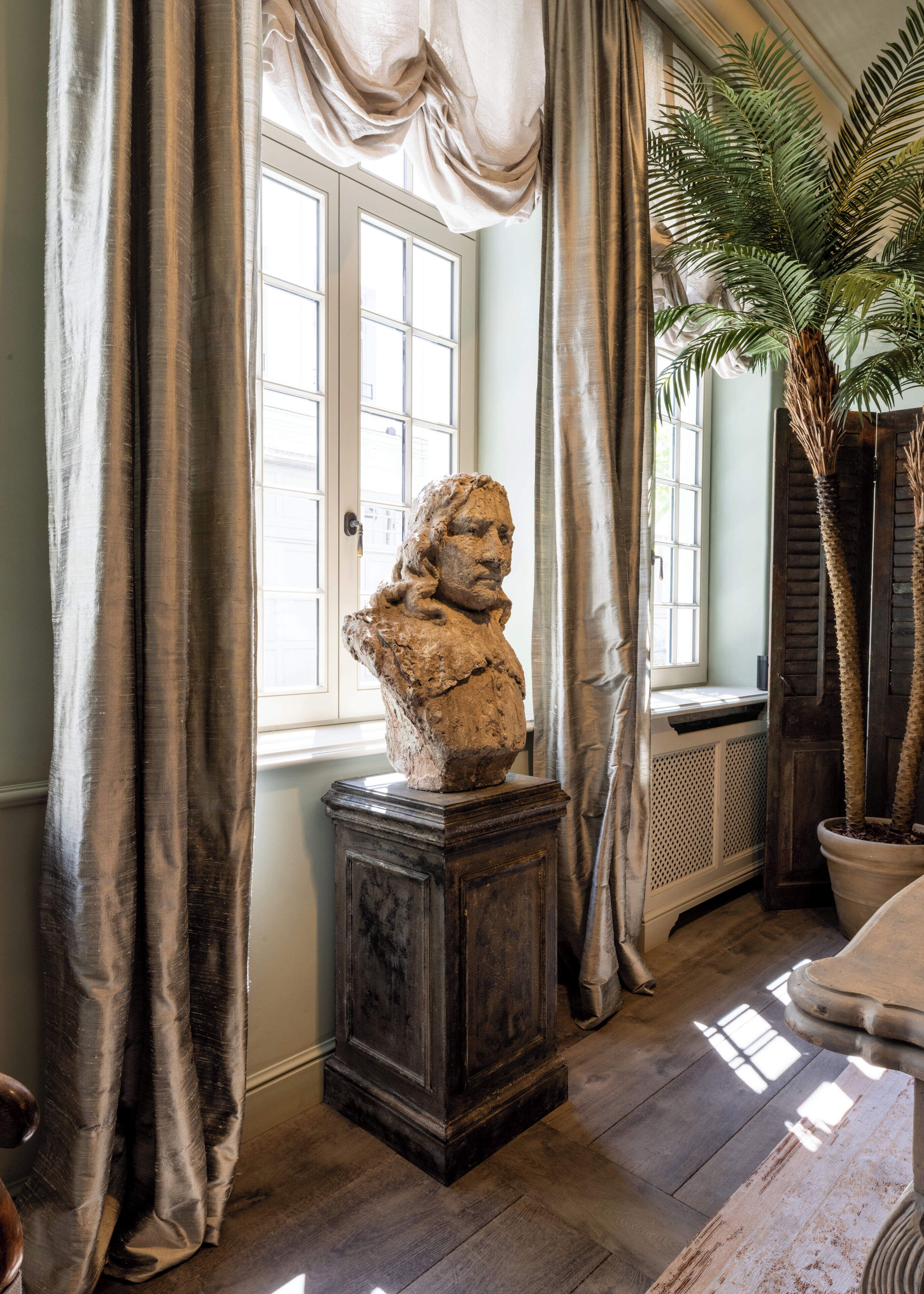 A room with a bust sculpture on a pedestal, a large window with curtains, and a potted palm plant.