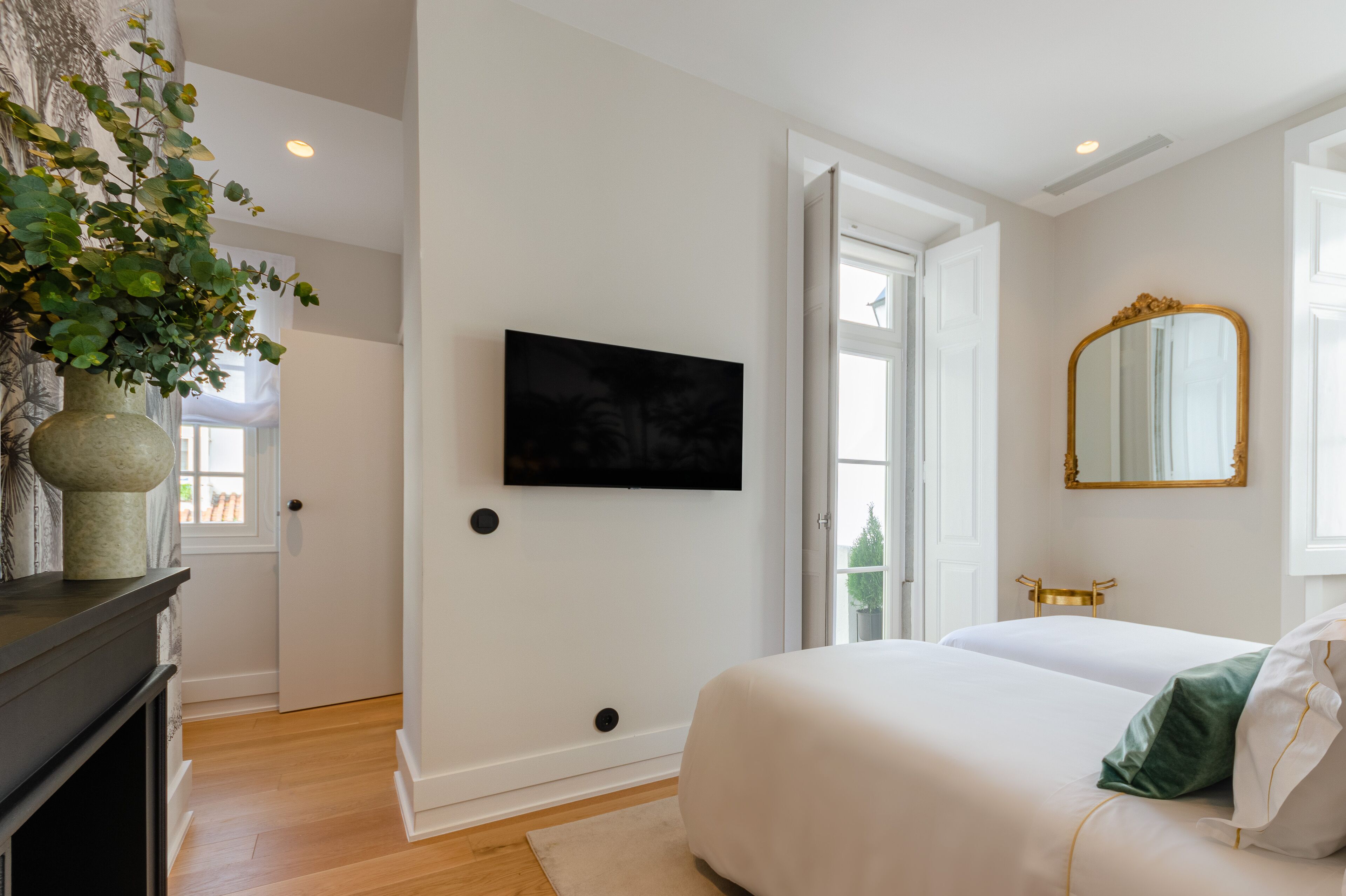 Un dormitorio con una cama, un televisor montado en la pared, un espejo y una puerta que da a otra habitación.
