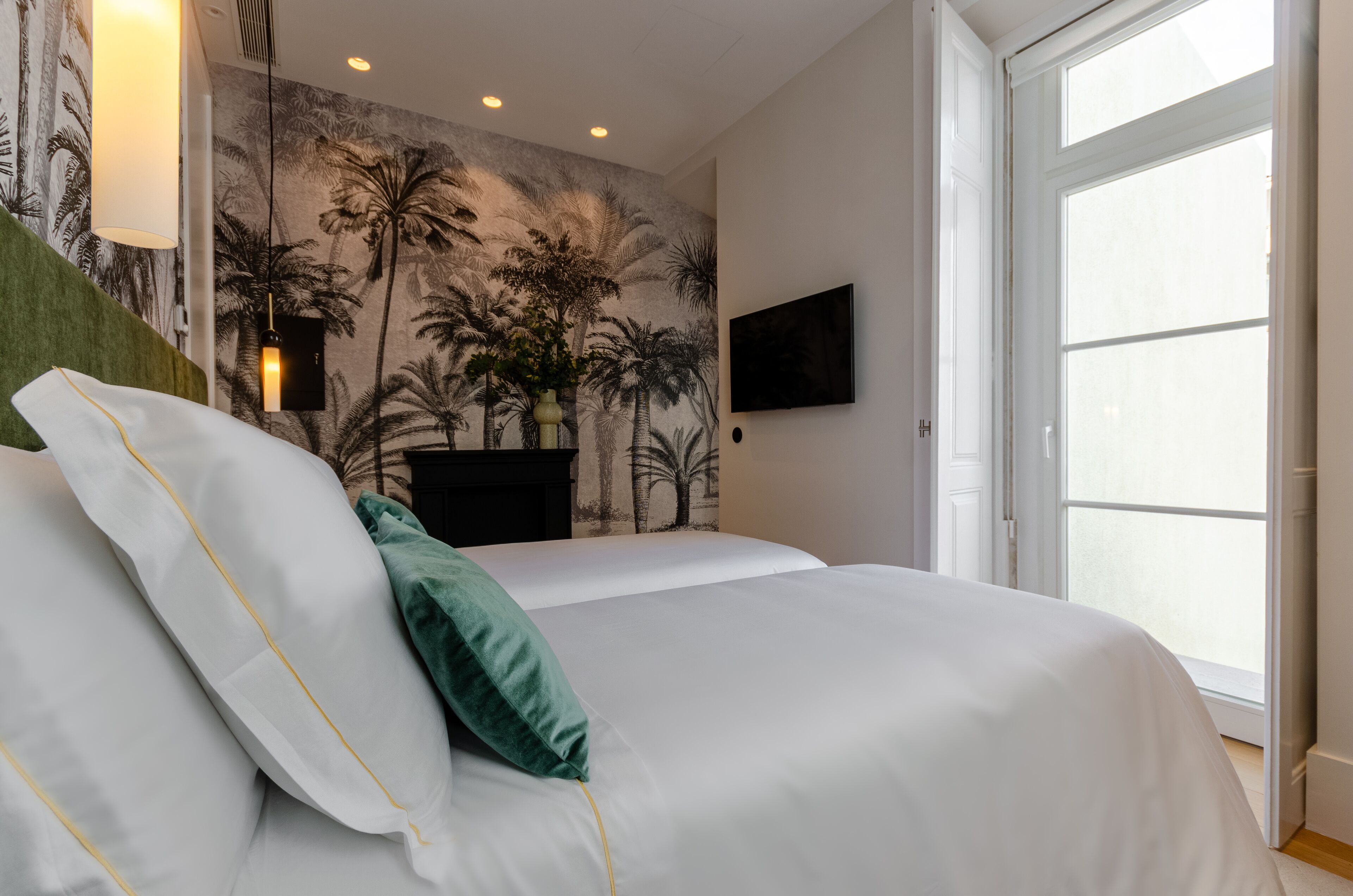 A neatly made bed with white and green pillows, a wall mural of palm trees, a mounted TV, and a large window.
