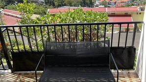 A balcony with a black metal bench, a small table with a bowl of fruit, and a water bottle. The balcony overlooks a coastal area with boats and buildings. - Maison Bel bonjou (Le Marin)