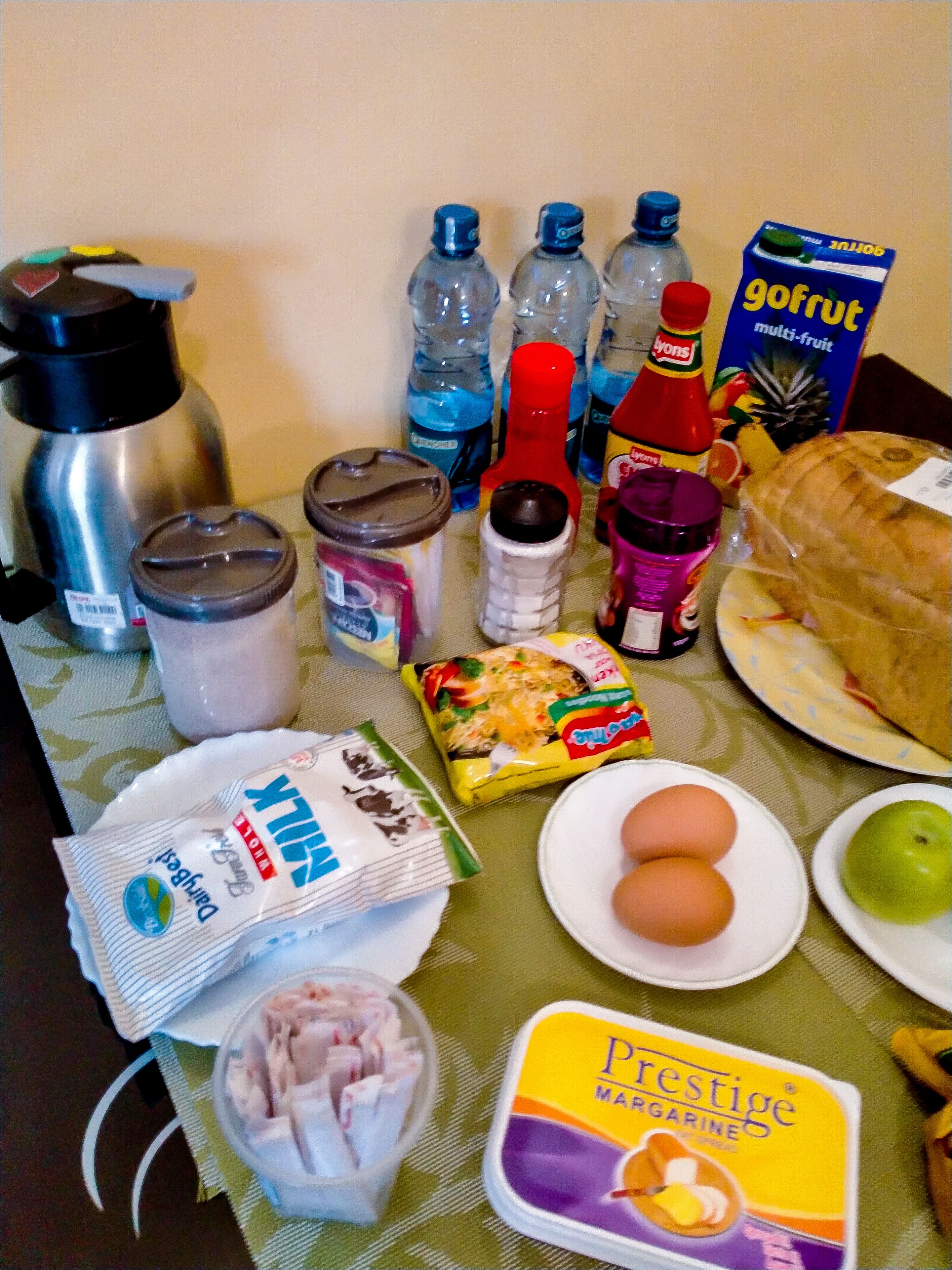 A table with various food items and condiments, including a thermos, water bottles, a bread roll, eggs, a green apple, and a box of margarine.