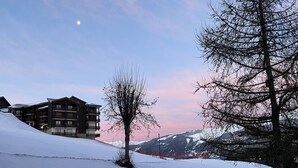 Paisaje nevado con un edificio que tiene balcones y un árbol en primer plano.