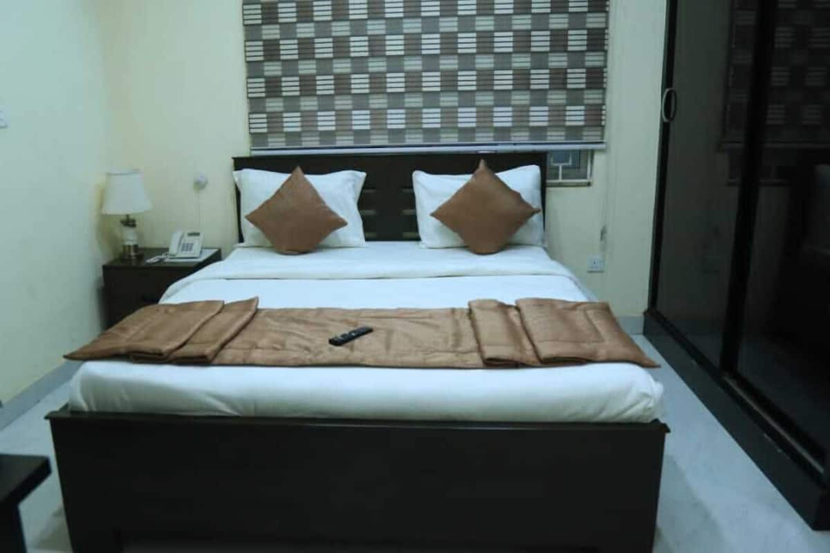 A hotel room with a bed, a bedside table with a lamp, a phone, and a window with blinds.