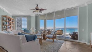 A spacious living room with a white sofa, blue cushions, a wooden bookshelf, and a view of the ocean through large sliding glass doors.
