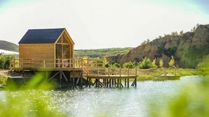 A wooden structure with a sloped roof on stilts over water, surrounded by greenery and hills. - Dahua Jianguo Hotel (Zhuolu)
