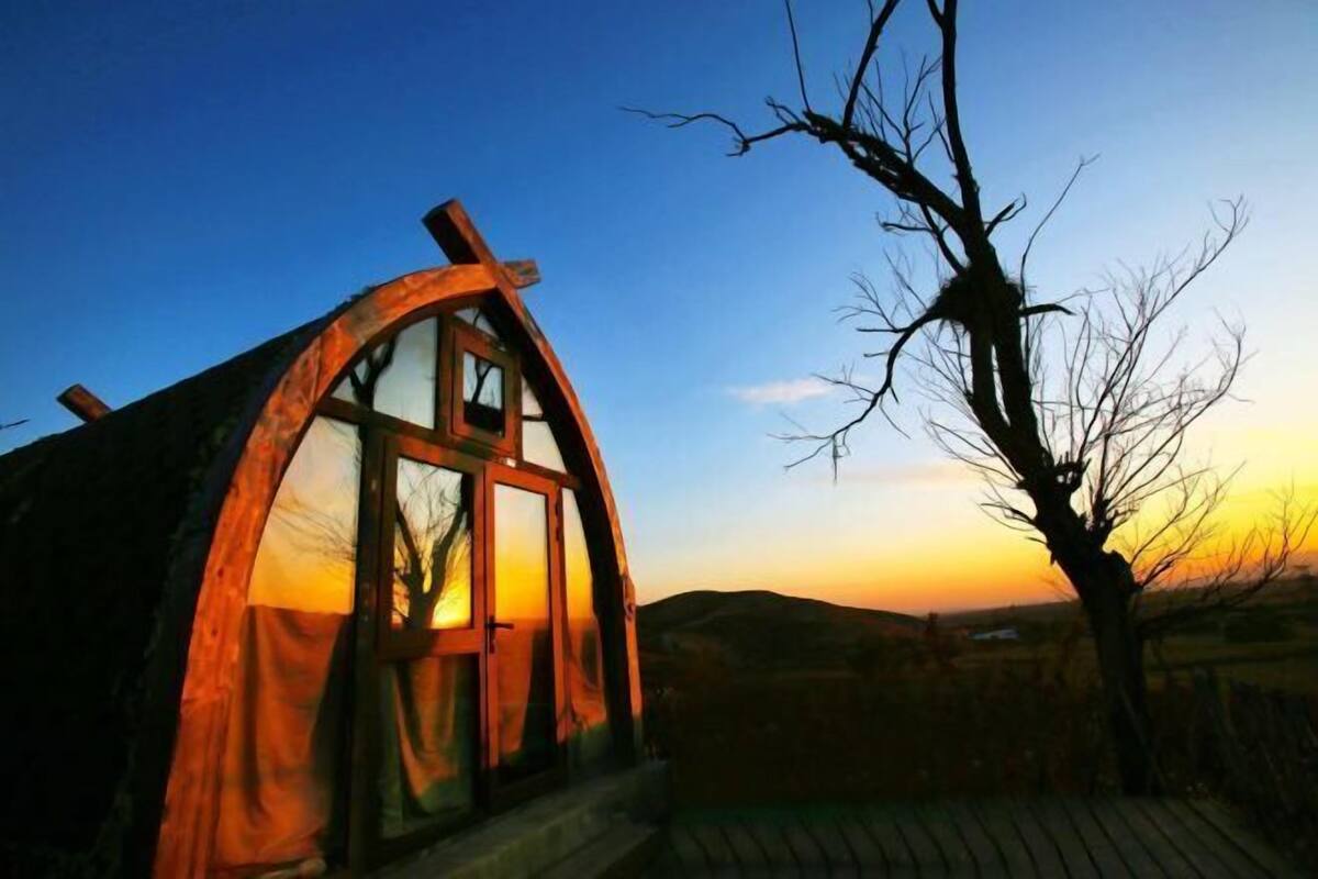 A rustic cabin with a large window, a wooden deck, and a bare tree in the foreground.