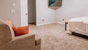 Ett sovrum med ett beige mattgolv, en säng med ett vitt överkast, ett nattduksbord i trä, en beige soffa med en orange kudde, en vit dörr och en takfläkt.