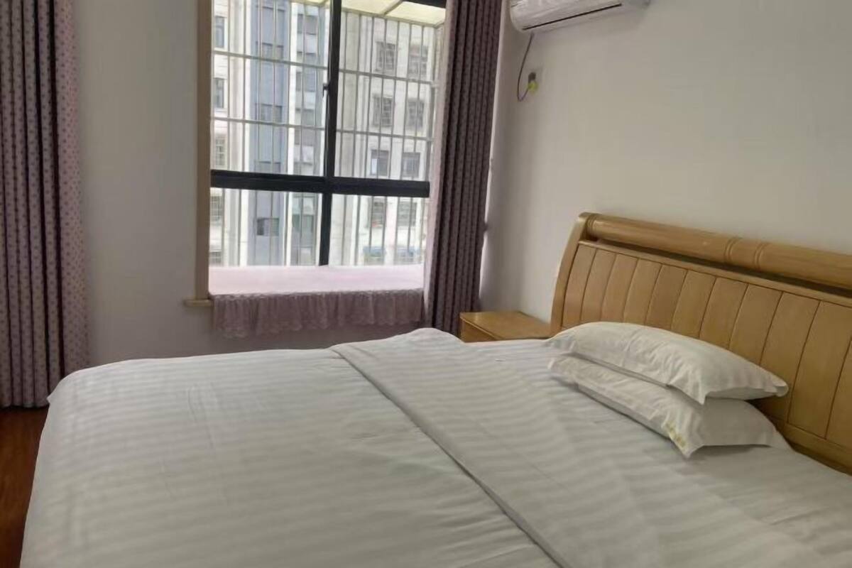 A hotel room with a bed, a window with a view of buildings, a bedside table, and an air conditioning unit.