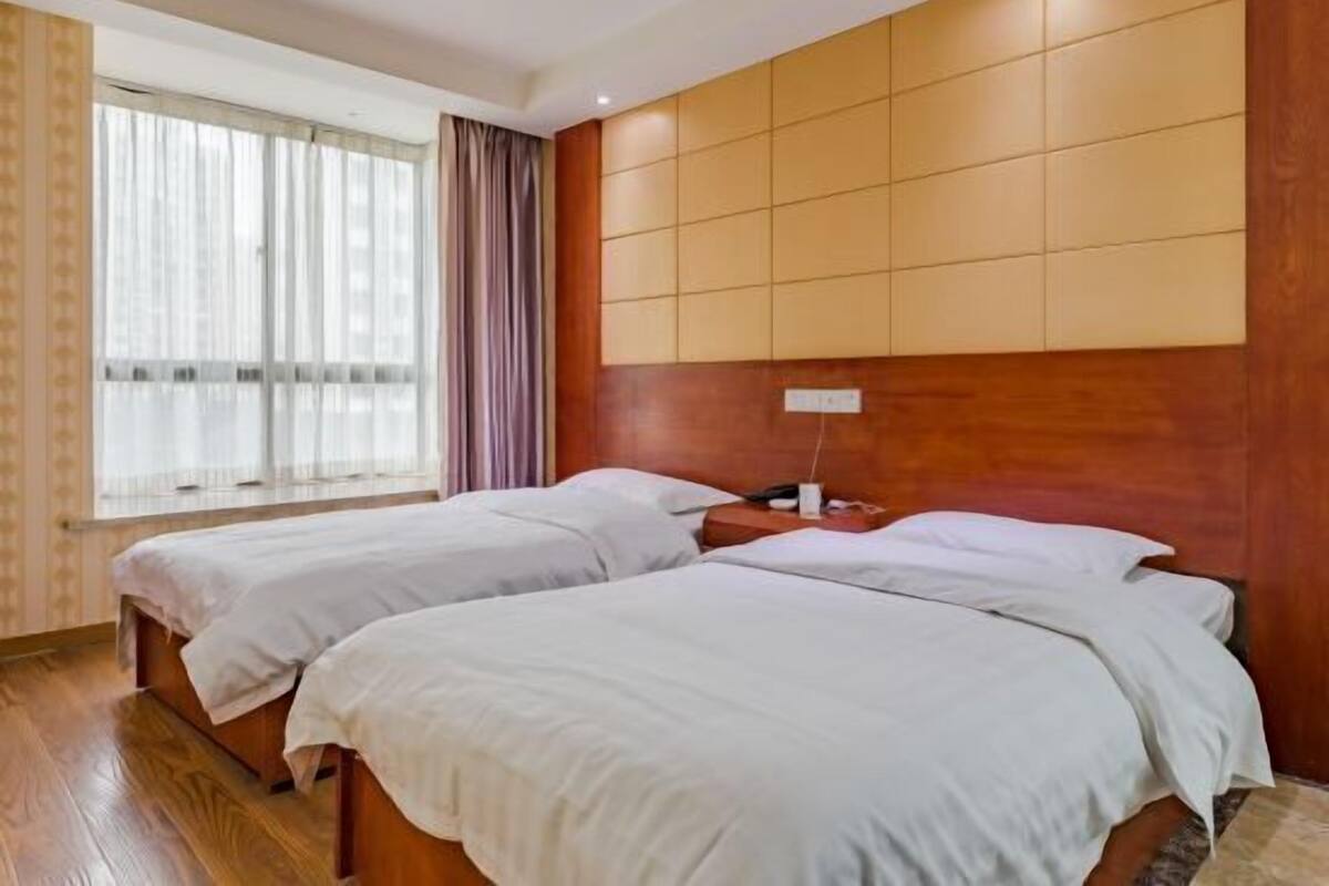 A hotel room with two beds, wooden flooring, a large window with curtains, and a wooden headboard with a panel design.