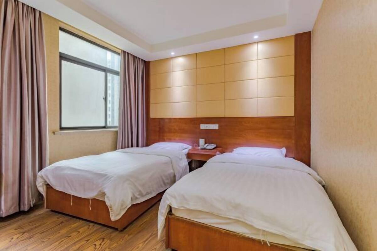 A hotel room with two beds, a wooden headboard, a window with curtains, and a bedside table with a phone.
