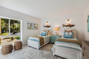A bedroom with two beds, a window with a view of trees and parked cars, and wall decorations including surfboards and a fish.