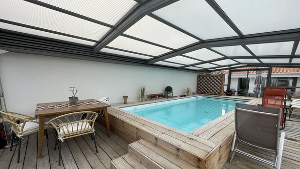 A covered outdoor swimming pool with wooden deck, a table with chairs, and a lounge chair.