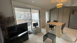 A modern living room with a flat-screen TV, a glass coffee table, a wooden dining table with chairs, and a kitchen area with a refrigerator.