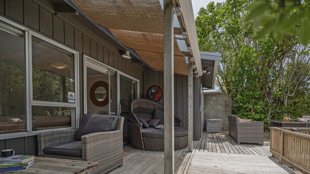 A covered outdoor deck with wicker furniture, a round table, and a wooden bench. - Kon Tiki Retreat - Whiritoa Beach Holiday Home (Whiritoa)