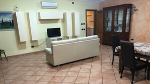A living room with a white sofa, a TV, and a dining table with chairs. - Cozy and independent apartment with garden! (Borgo A Buggiano)