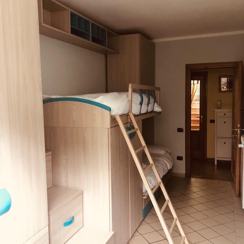 Un lit superposé avec une échelle, un petit bureau et un placard dans une pièce dont le sol est recouvert de carreaux.