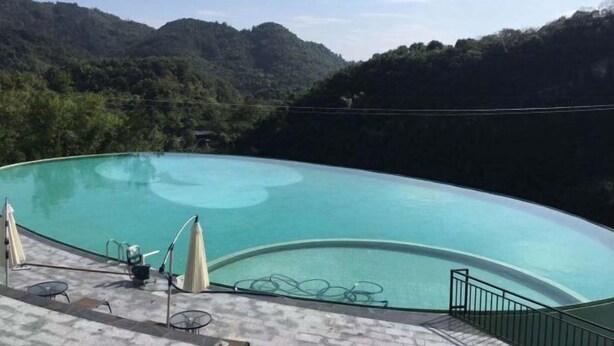 A curved infinity pool with lounge chairs and umbrellas, surrounded by lush greenery and hills. - Cliff Hotel (Luocheng)