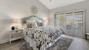 A bedroom with a large bed, a ceiling fan, a decorative wall piece, and a window with blinds.