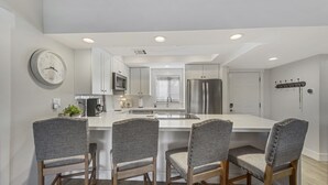 A modern kitchen with white cabinetry, a central island with bar stools, a stainless steel refrigerator, and a microwave.