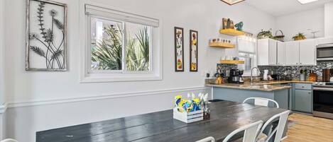 A modern kitchen with white cabinets, a dark wooden dining table, and a window with a view of palm trees.