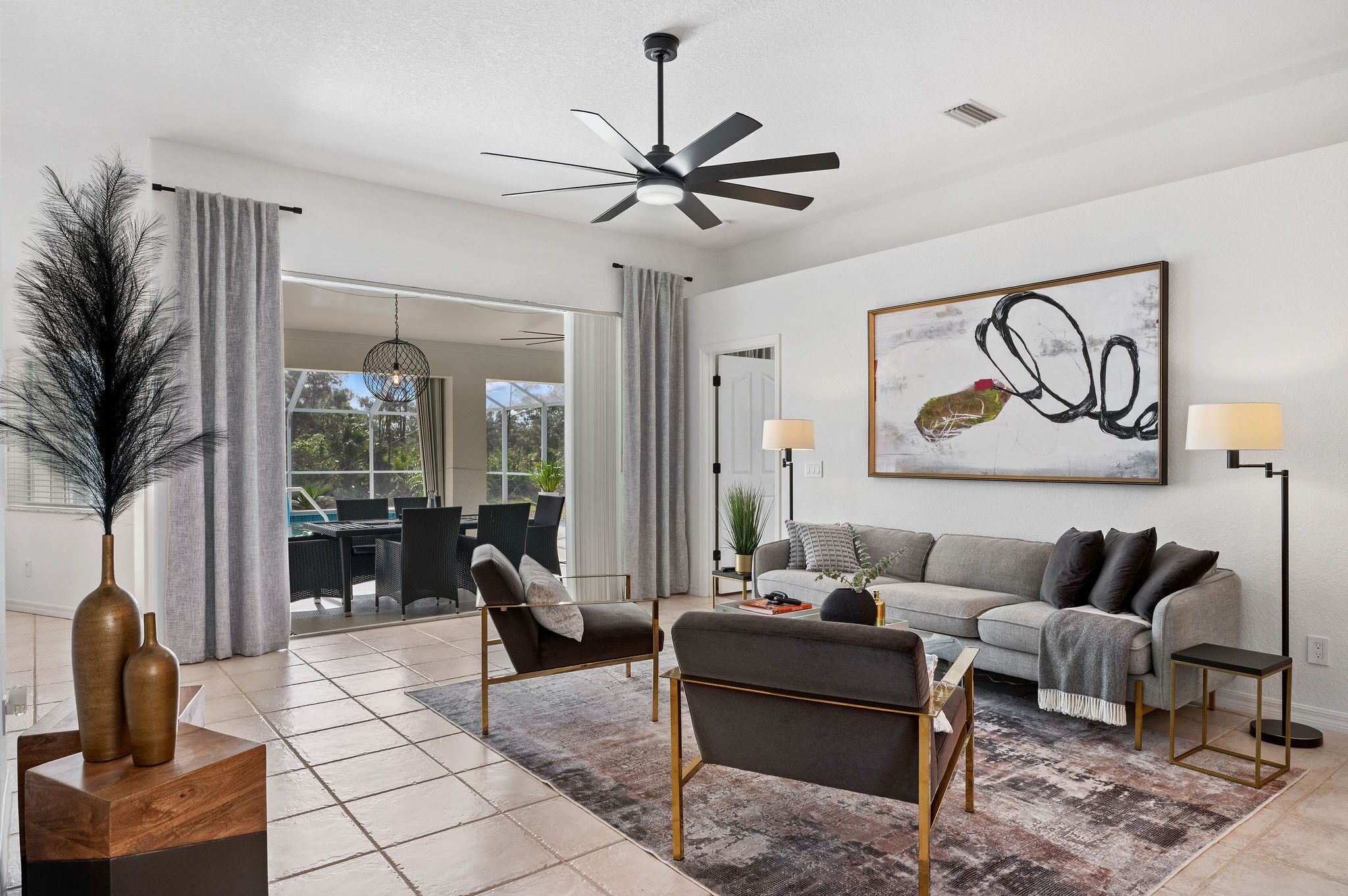 A modern living room with a grey sofa, two armchairs, a coffee table, and a floor lamp. There is a ceiling fan, a painting on the wall, and a view of the outdoors through large windows.