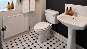 Une salle de bain avec des toilettes, un lavabo et un miroir. Il y a une fenêtre avec des stores, une étagère avec une plante et des serviettes, et un sol carrelé.