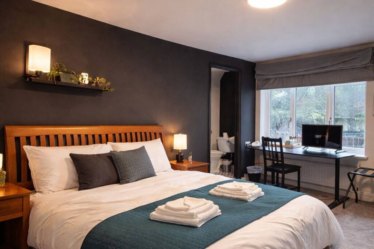 Quarto de hotel com uma cama grande, mesas de cabeceira, escrivaninha com cadeira, janela e televisão.