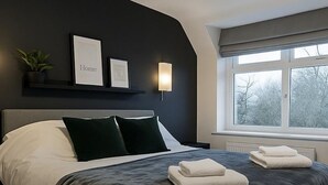 Une chambre moderne avec une tête de lit sombre, un lit avec une literie blanche et foncée, une table de chevet avec une lampe et une fenêtre donnant sur des arbres.