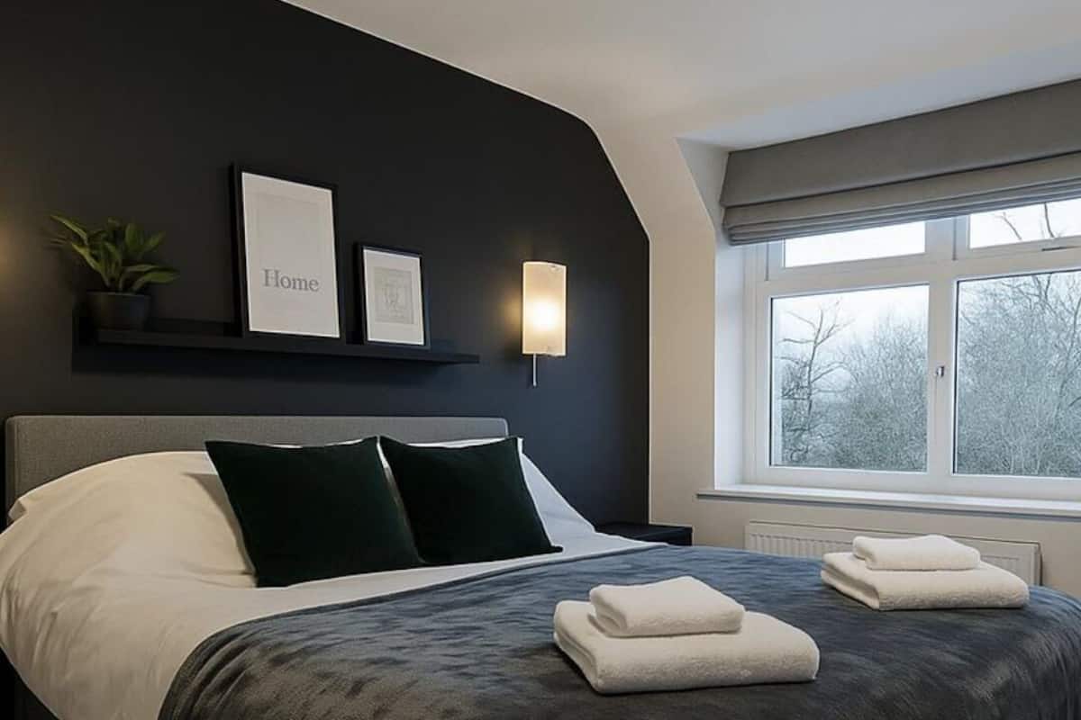 Quarto moderno com cabeceira escura, cama com roupa de cama branca e escura, mesa de cabeceira com abajur e janela com vista para árvores.