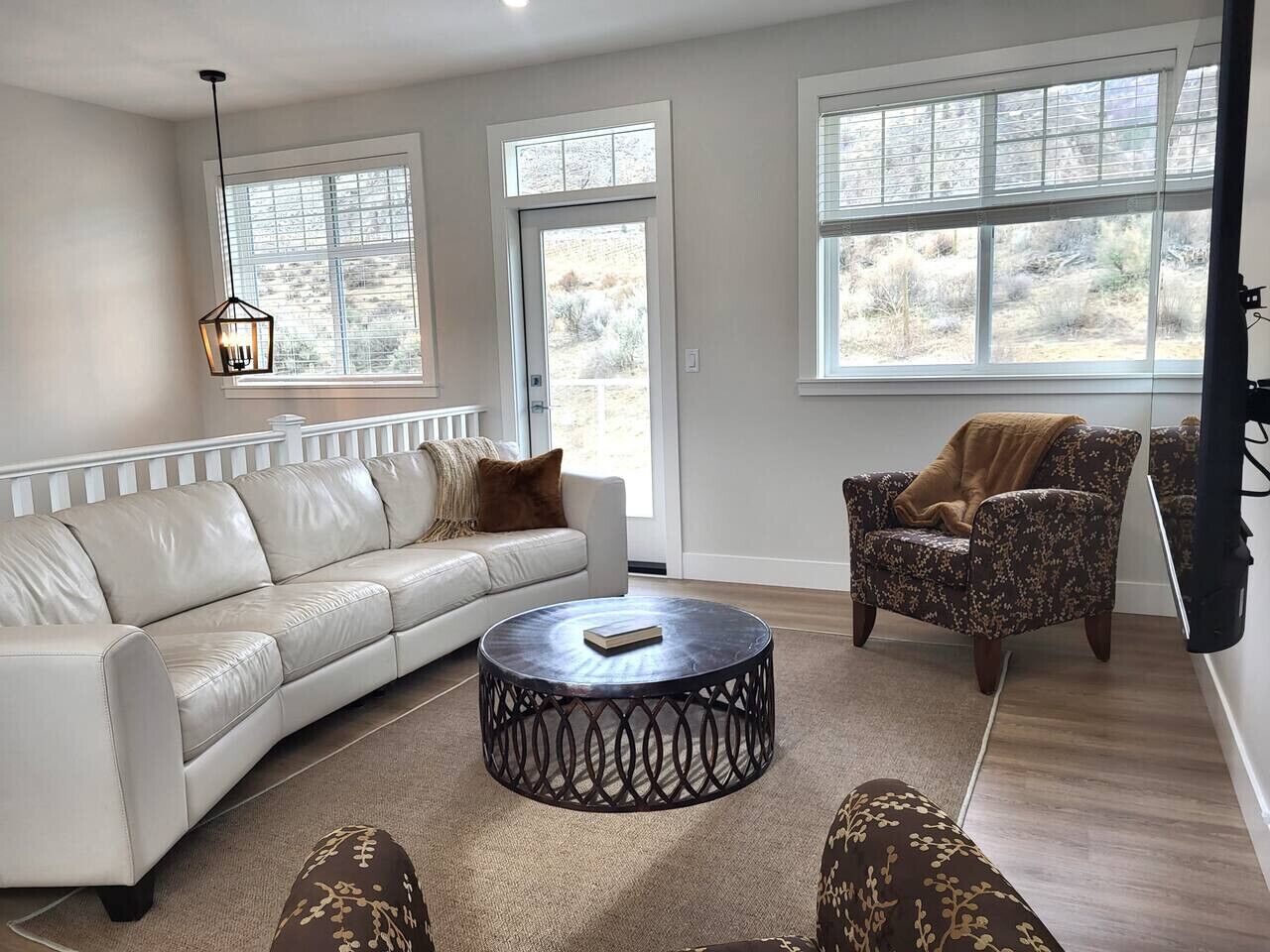 Una sala de estar con un sofá de cuero blanco, una mesa redonda, un sillón estampado y ventanas grandes.