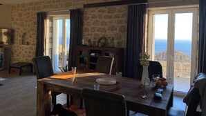 Un'area pranzo con parete in pietra, tavolo di legno e sedie. Si apre una vista sul mare attraverso ampie finestre.