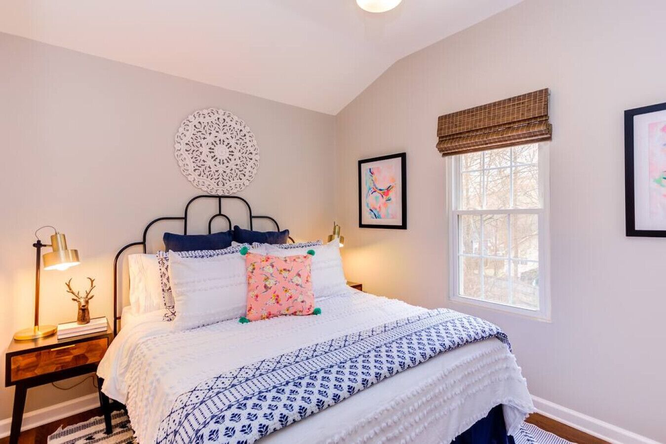 A bedroom with a bed, bedside tables, a lamp, a decorative wall piece, a window with blinds, and a framed picture on the wall.