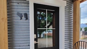 Un chalet rustique avec une façade en métal ondulé, une porte noire dotée d’un cadre blanc, un petit porche avec une chaise et un paillasson.