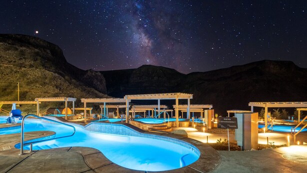 Un espace de baignade nocturne comprenant plusieurs piscines à ciel ouvert, sous un ciel étoilé, entouré de structures en bois et dominé par un paysage montagneux.