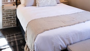 A bedroom with a brick wall, a bed with white bedding, and a large artwork of a cow's head above the bed.