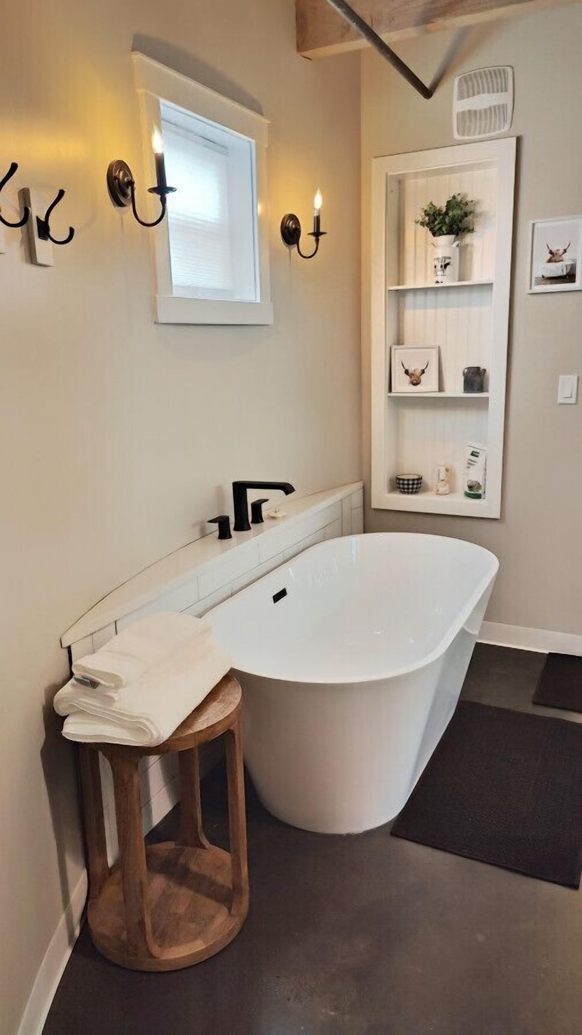 Un baño moderno con una bañera independiente, un taburete de madera, un tocador blanco y un estante de pared con adornos.