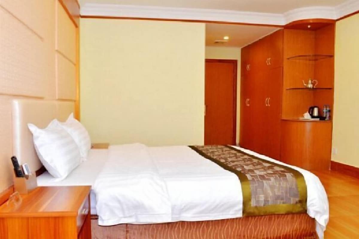 Une chambre d’hôtel avec un lit bien fait, une table de chevet en bois, une armoire et une petite étagère sur laquelle se trouve une bouilloire.