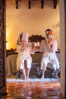 Dos personas vestidas de blanco sentadas en una cama con cabecera de madera, sosteniendo tazas y un libro.