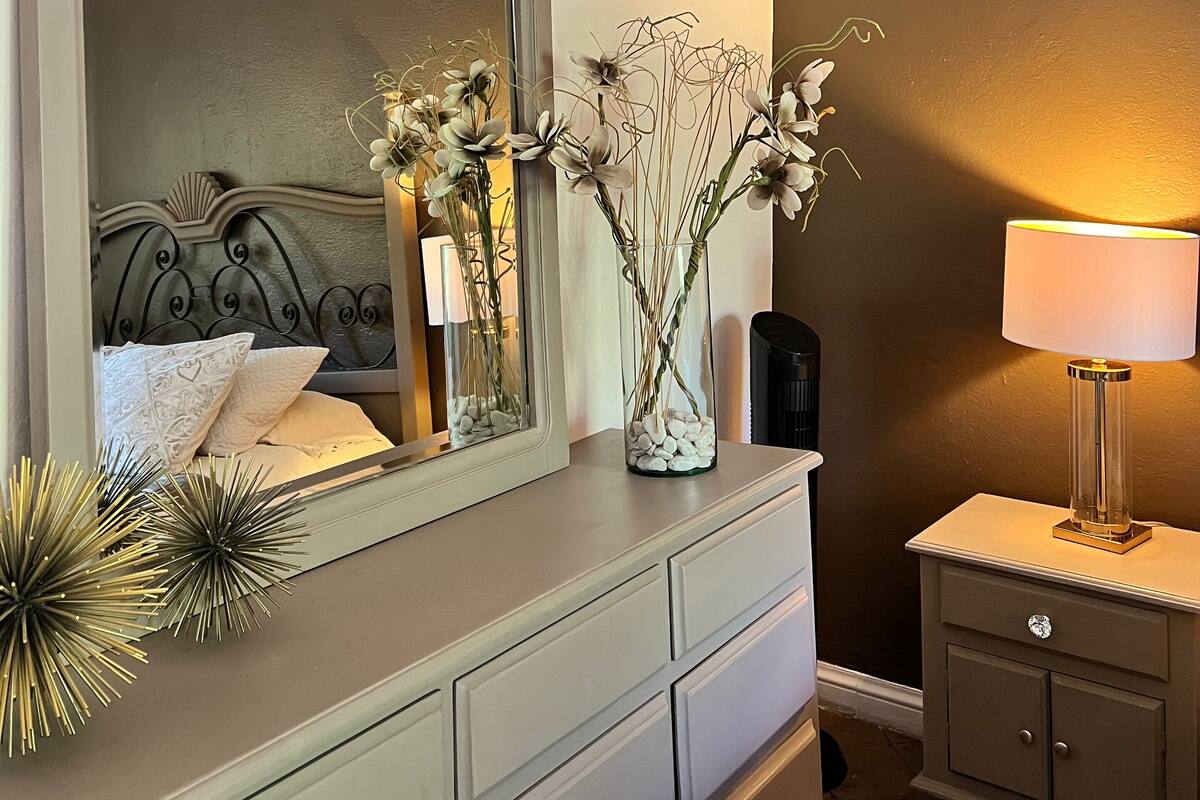 A bedroom with a vanity, a mirror, a bedside table with a lamp, and a decorative plant.