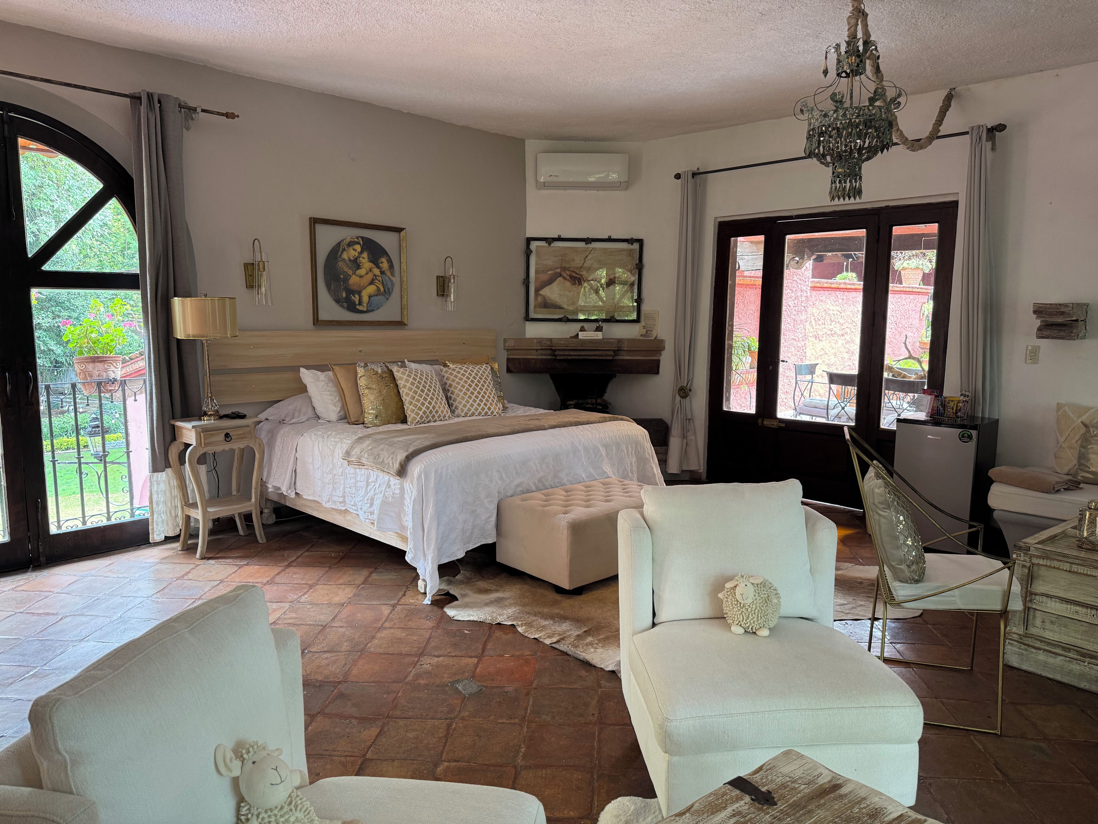 Une chambre à coucher comprenant un lit, des tables de chevet, une commode, une chaise, un lustre et une vue sur l’extérieur, visible à travers une porte.