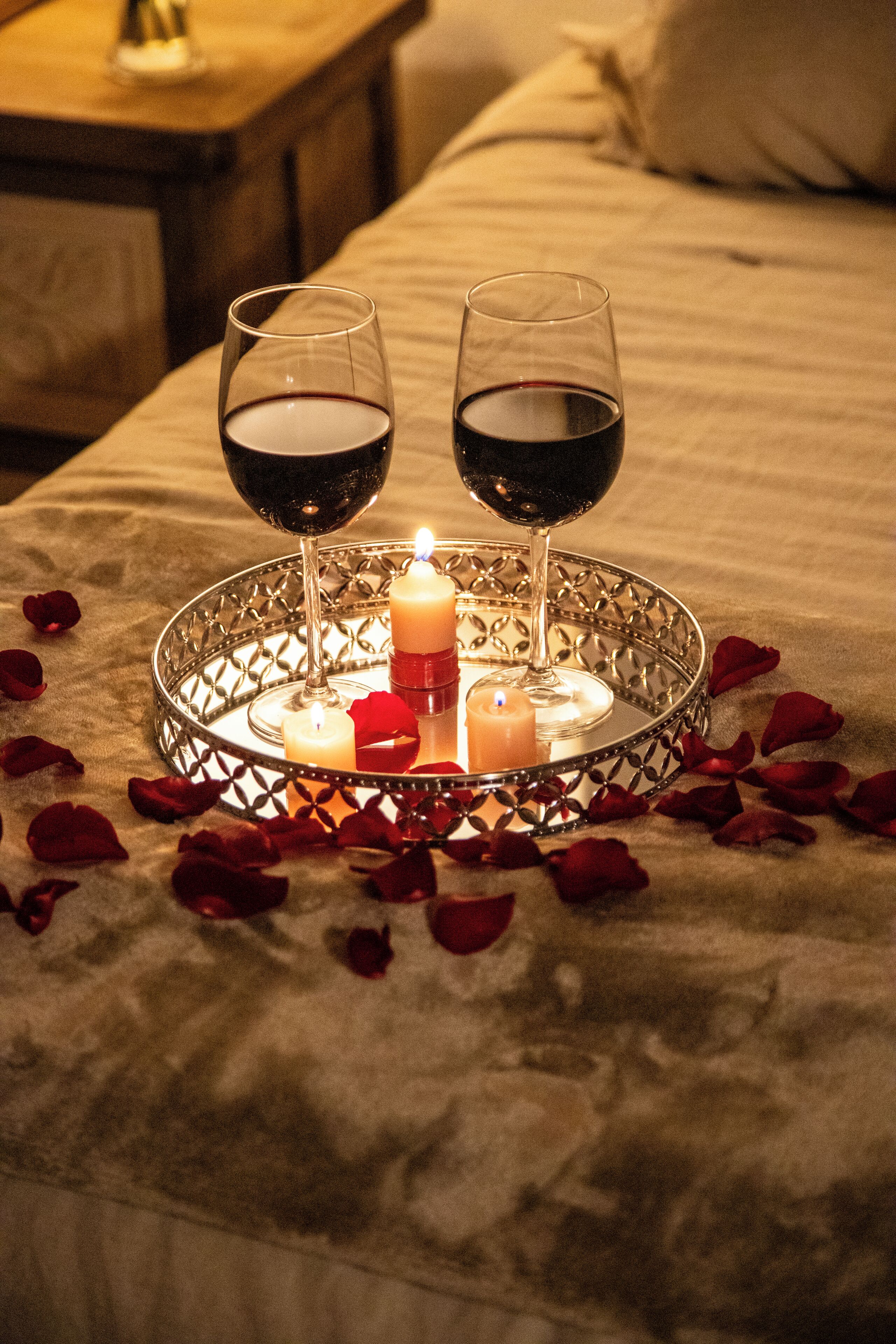 A tray with two glasses of red wine, a candle, and rose petals on a bed.