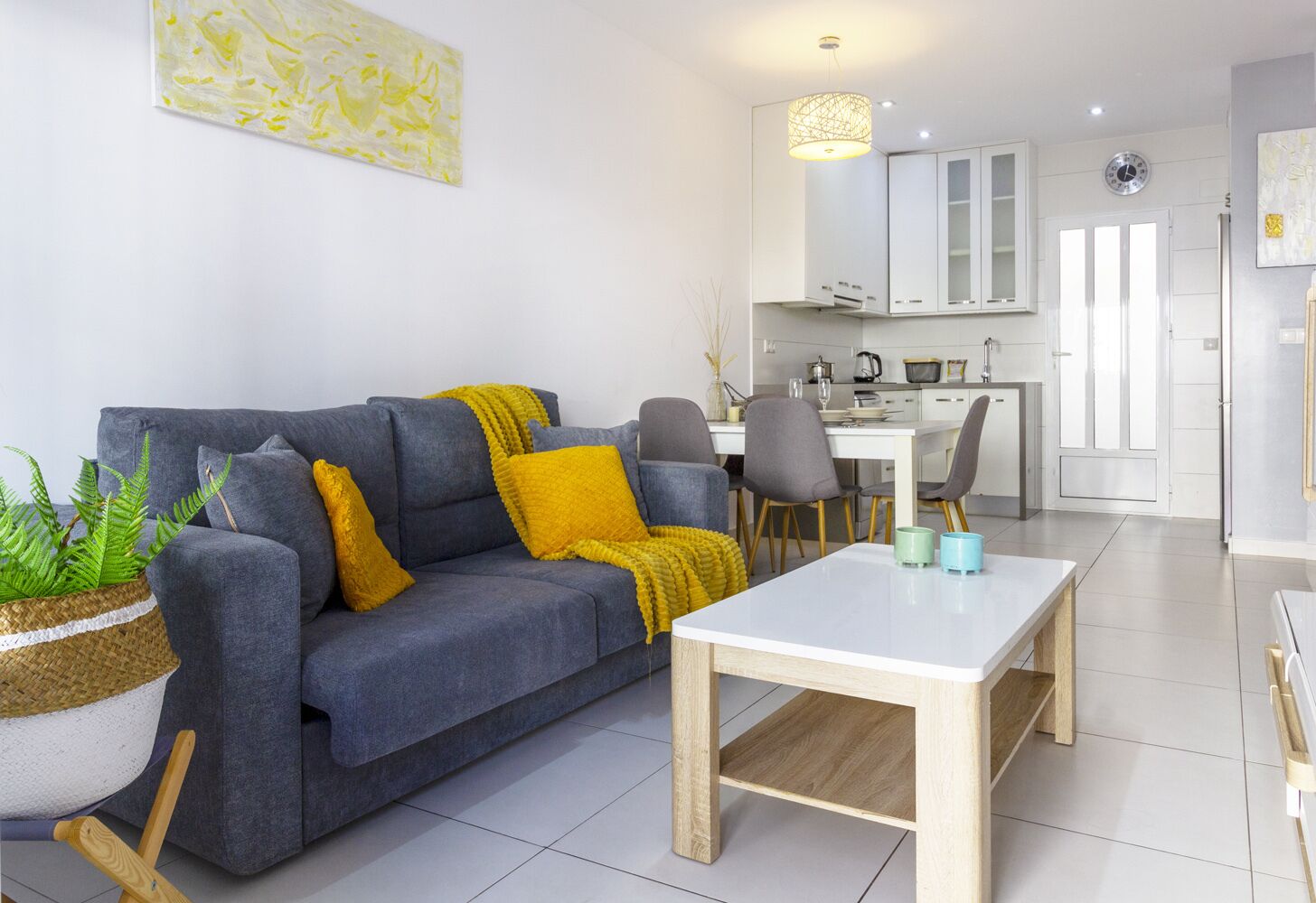 Una sala moderna con un sofá gris, almohadones amarillos, una mesa de centro blanca y un comedor con una mesa y sillas blancas.