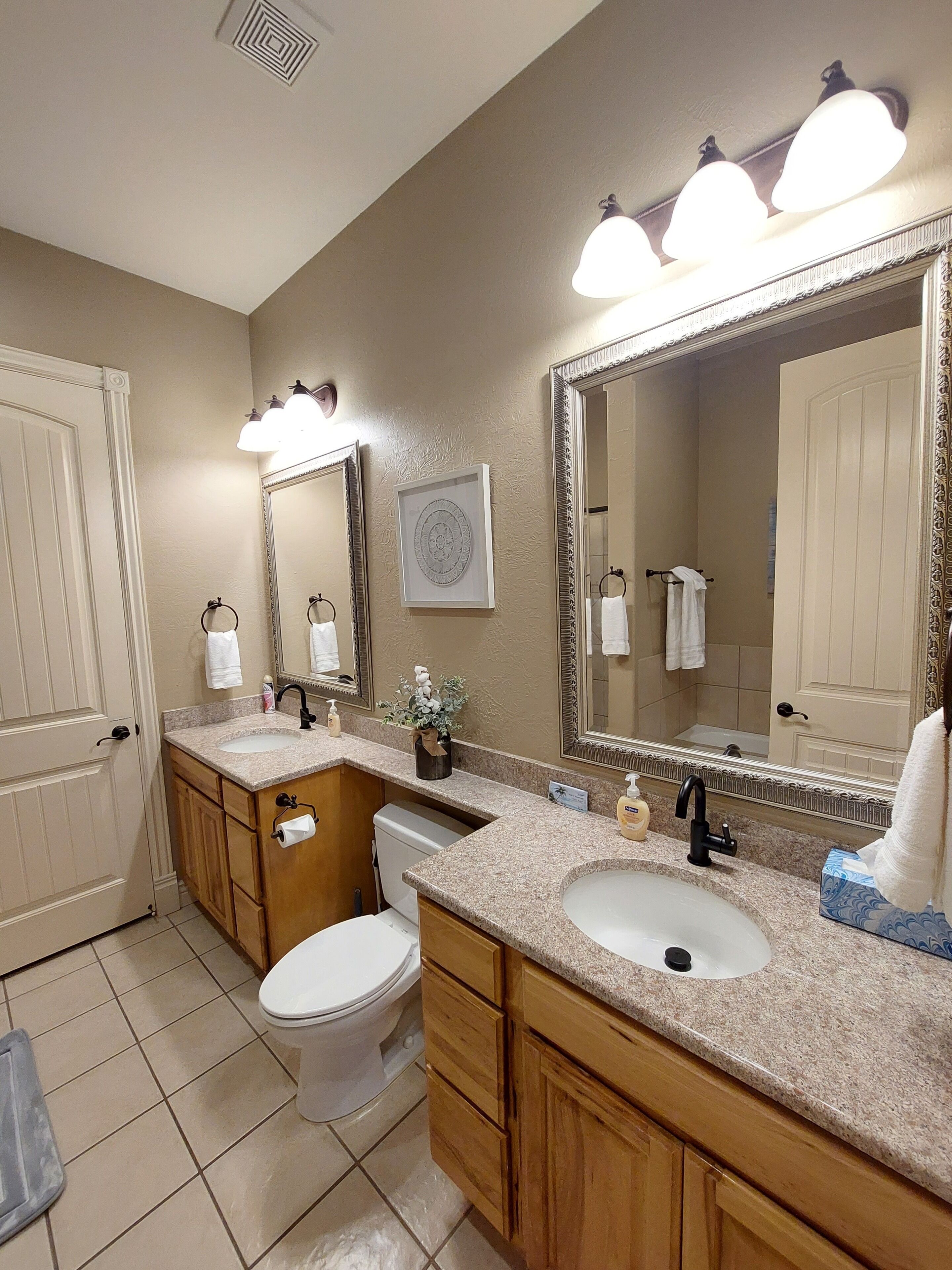 A bathroom with two sinks, a toilet, and a large mirror. There are towels hanging on the wall and a framed picture above the sink.