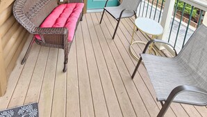 Eine Holzterrasse mit einem Korbsessel und zwei Stühlen, einer türkisblauen Tür und Blick auf eine grüne Landschaft.
